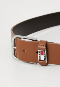 Brun lærrem med en sølvfarget metallspenne, som har et Tommy Jeans-logo. Jevnlige teksturer, med hull for justering.
