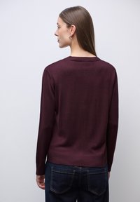 Pullover bordeaux con scollo rotondo, maniche lunghe, lavorazione a maglia testurizzata e orlo a costine, indossato sopra jeans in denim scuro.