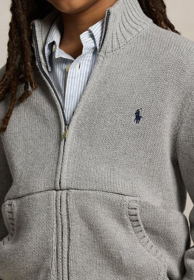 Person, die einen grauen Strickpullover mit Reißverschluss, geripptem Kragen und Taschen trägt, über einem blau-weiß gestreiften Hemd mit Knöpfen.