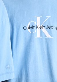 Vaaleansininen puuvillapaita, jossa on suuri valkoinen "Calvin Klein Jeans" -logo ja pieni brodeerattu "ck" hihassa. Sileä pinta.
