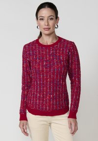 Pull en tricot rouge avec un motif en grille comportant des accents bleus et blancs. Encolure, poignets et ourlet côtelés. Manches longues.