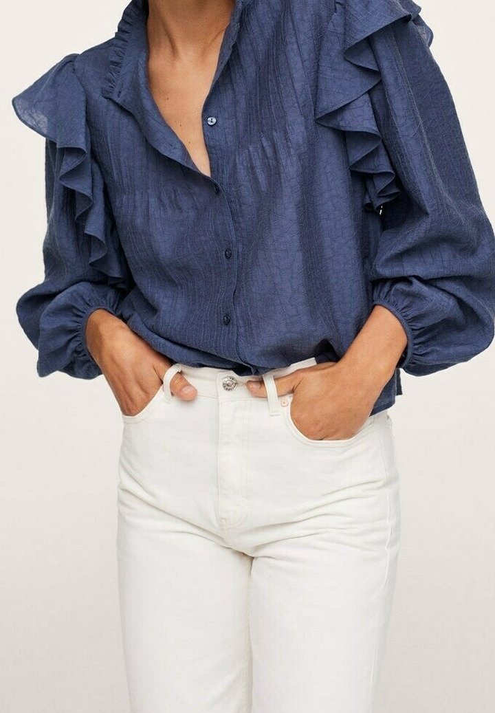 Femme portant un chemisier bleu marine à manches longues avec des volants, rentré dans un pantalon blanc taille haute, les mains dans les poches.