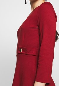 Czerwona sukienka z długim rękawem, uszyta z miękkiego materiału, o dopasowanym kroju, z dekoltem w kształcie litery V oraz przednią kieszenią ozdobioną złotym guzikiem.