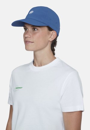 Frau mit geflochtenem Haar, die eine blaue Kappe und ein weißes Mammut-T-Shirt trägt, schaut nach links vor einem einfarbigen Hintergrund.