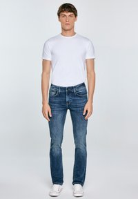 Jeans de mezclilla azul de corte Slim fit con un lavado desgastado, combinados con una camiseta blanca de cuello redondo. Zapatillas blancas completan el conjunto.