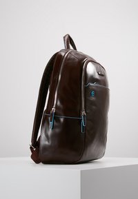 Mochila de cuero marrón con dos cremalleras frontales, detalles en azul, un acabado texturizado y un asa en la parte superior. Diseñada para la funcionalidad y el estilo.