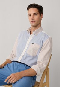 Korte mouwen button-down overhemd in lichtblauw en beige stof, met een borstzak, zachte textuur en contrasterende kleuren.