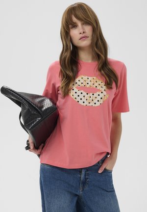 Kvinde med langt brunt hår iført en koralfarvet t-shirt med et guld- og polkaprikket læbemotiv, blå jeans, holder en sort flettet taske.