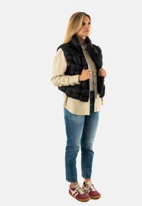 Chaleco acolchado negro sobre una camisa beige, combinado con jeans azules y zapatillas burdeos. El chaleco tiene un cuello alto y no presenta herrajes visibles.