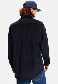 Dark corduroy shirt with a pointed collar, buttoned cuffs, and a back yoke design; paired with blue jeans and a navy cap.