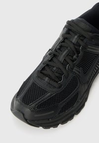 Chaussure de sport noire avec une tige en maille texturée et en matière synthétique, dotée d'une languette rembourrée, de lacets plats et d'une semelle en caoutchouc épaisse.