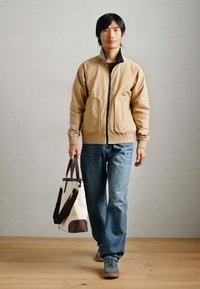 Μαύρο bomber μπουφάν με φερμουάρ, δύο τσέπες; μπλε τζιν; γκρι αθλητικά παπούτσια; κρατώντας μια κρέμα και καφέ τσάντα. Ξύλινο δάπεδο, ουδέτερο φόντο.