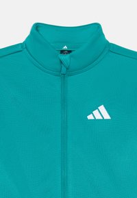 T türkiz sportdzseki magas gallérral, teljes cipzárral és fehér Adidas logóval. Simas, könnyű anyagból készült, struktúrált fazonnal.
