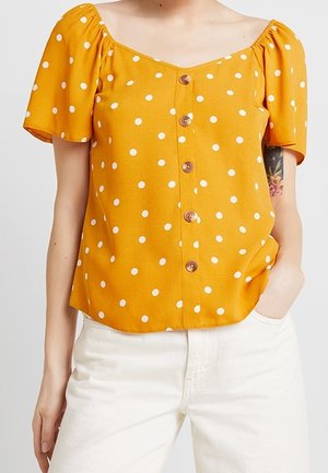 Femme portant un chemisier jaune à manches courtes avec des pois blancs et des boutons sur le devant, associé à un pantalon blanc.