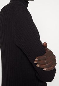 Maglione a collo alto nero a costine con maniche lunghe. Primo piano del dettaglio del braccio, che mostra il punto tessuto e il design aderente.