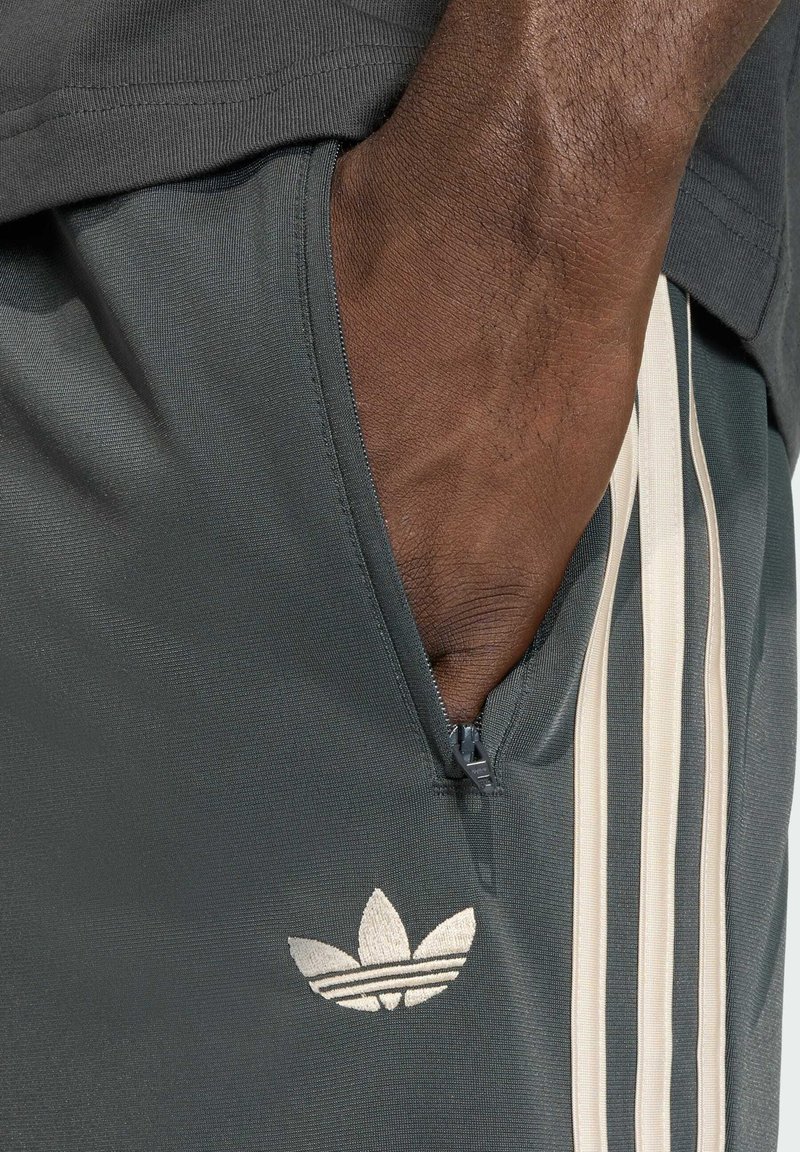 Graue Sportshorts mit einem Reißverschlusstaschen, versehen mit einem silbernen Adidas-Logo und cremefarbenen Streifen an der Seite. Glatte Textur.