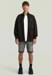 Veste zippée noire avec deux poches avant, portée sur un t-shirt blanc et un short gris déchiré. Le mannequin porte des baskets noires et des chaussettes.