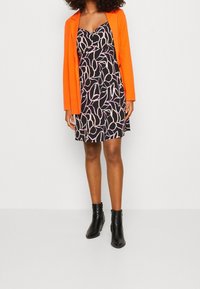 Robe noire avec des motifs de feuilles roses et blanches, portée sous un blazer orange. Des bottines noires agrémentent l'ensemble.