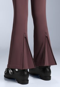 Strakke skibroek in donker bordeauxrode stof, met zijritsen bij de boorden en gecombineerd met zwarte skischoenen.