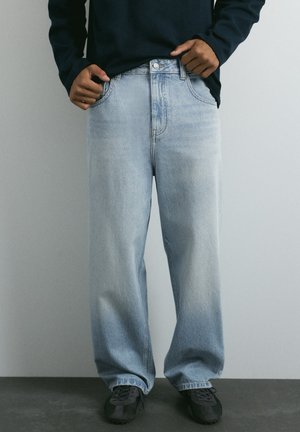 Personne portant un jean bleu clair ample, des baskets noires et un haut sombre à manches longues avec les mains près des poches à la taille.