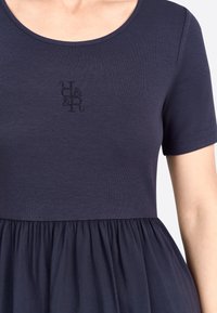 Robe bleu marine avec un haut côtelé et des manches courtes, un bas froncé, et un logo brodé sur la poitrine. Texture douce dans l'ensemble.