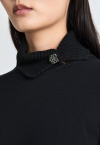 Schwarzer Pullover mit einem gerippten Kragen und einer dekorativen quadratischen Brosche aus metallischen und kristallinen Akzenten auf der linken Seite.
