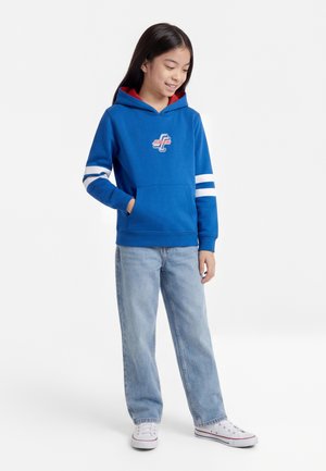 Girl wearing a blue hoodie with white stripes and logo, light blue jeans, and white sneakers, standing against a plain background.