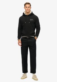 Sudadera negra con capucha y bolsillo frontal, superpuesta a una sudadera gris. Combinada con pantalones negros y zapatillas beige, con un reloj.