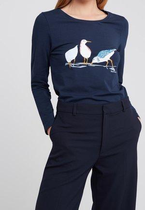 Kobieta w granatowej koszuli z długim rękawem z trzema ilustrowanymi ptakami stojącymi na gałęzi, w połączeniu z pasującymi granatowymi spodniami.