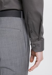 Szara koszula z fakturowanej tkaniny, zapięta na dwa guziki na mankietach, połączona z dopasowanymi szarymi spodniami i czarnym paskiem widocznym w talii.