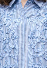 Μπλε και άσπρο ριγέ πουκάμισο με κουμπιά, με ανασηκωμένες, βολβοειδείς λεπτομέρειες ύφασματος που δημιουργούν αφηρημένα μοτίβα στο μπροστινό μέρος.