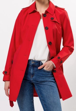 Mujer con un abrigo rojo brillante abotonado sobre una camiseta blanca y jeans azules, con una mano en el bolsillo, frente a un fondo liso.