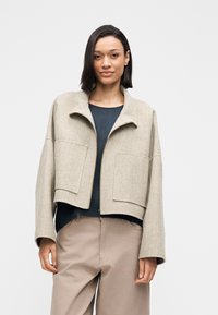 Beżowa krótka kurtka z wysokim kołnierzem, dwoma dużymi przednimi kieszeniami i szerokimi rękawami, wykonana z teksturowanej tkaniny. Noszona na ciemnym topie.