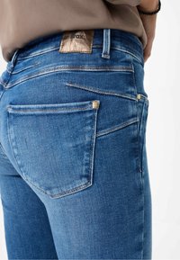 Jeans en denim bleu avec une finition texturée, dotés d'un patch en cuir marron, de poches arrière et de détails de couture subtils.