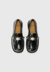 Schwarze Lederloafers mit glänzender Oberfläche, mit einer dekorativen silbernen Kette und Emblemdetail auf der Vorderseite. Strukturiertes Schuhsohle mit Nähakzenten.
