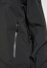 Section d'une veste imperméable noire montrant deux poches verticales zippées et un logo de marque brillant sur la manche supérieure.