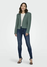 Groene, gestructureerde blazer met een cropped ontwerp, gecombineerd met een witte trui en donkere spijkerbroek. Model staat met zwarte hoge hakschoenen.