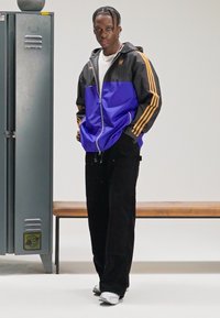 Μαύρο και μωβ αθλητικό μπουφάν με πορτοκαλί ρίγες, σε συνδυασμό με μαύρο παντελόνι με φαρδιά πόδια. Στο φόντο υπάρχει ένα ξύλινο παγκάκι και ένα ντουλάπι.