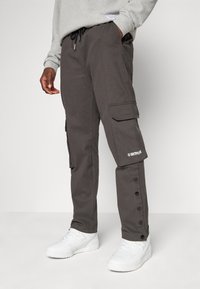 Grå cargo bukser med sidelommer, justerbar snor i taljen, trykknappslukking på sidene, og en logo på leggen. Brukt med hvite joggesko.
