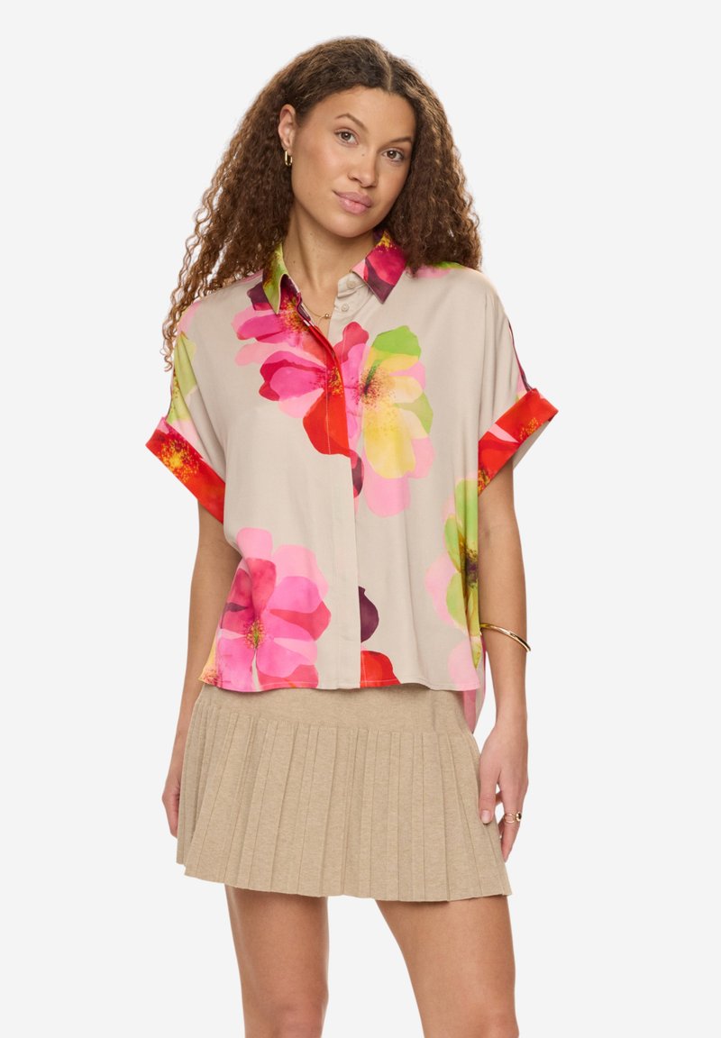 Femme aux cheveux bouclés portant une jupe plissée beige et une chemise à manches courtes boutonnée avec de grands motifs floraux roses et rouges.