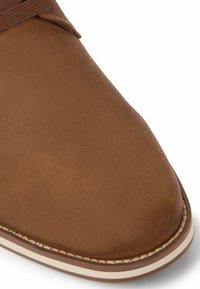 Chaussure en cuir marron au toucher lisse, à bout arrondi et avec des coutures blanches en accent. Design à lacets avec une légère brillance.