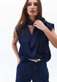 Námořnická modrá bezrukávová halenka s výstřihem do V a detailem šňůrky kolem krku, spárovaná s odpovídajícími kalhotami s vysokým pasem a světlým páskem.