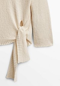 Pull en tricot crème avec un détail noué sur le côté. Présente une texture côtelée et des manches trois-quarts.