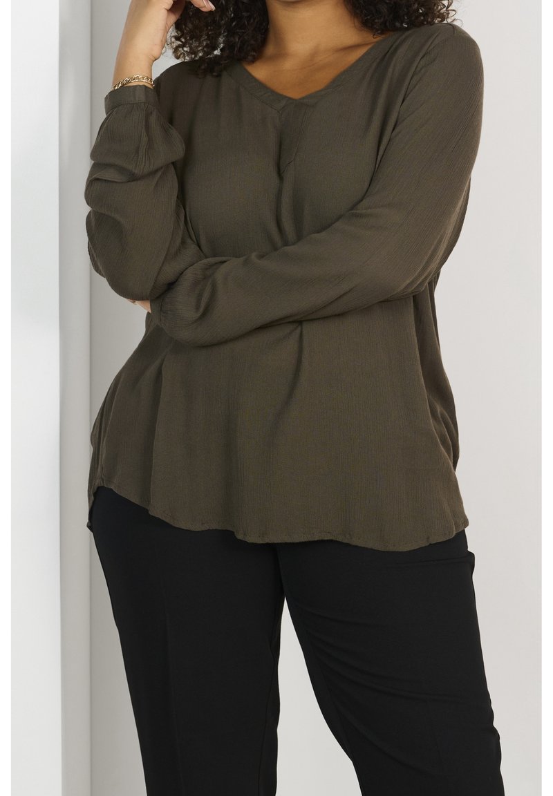 Blouse vert olive avec un col en V et de longues manches bouffantes. Fabriquée en tissu léger avec une coupe fluide et décontractée et un ourlet arrondi. Associée à un pantalon noir.