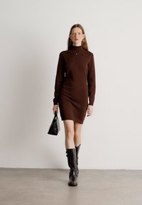 Vestido marrón ajustado de manga larga hecho de un tejido de punto texturizado. La modelo sostiene un pequeño bolso negro y lleva botas negras hasta la rodilla.