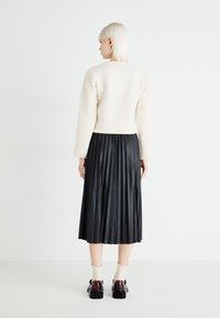 Krämfärgad fluffig kort jacka i kombination med en svart veckad midikjol. Modellen bär bruna skor och krämfärgade ankelsockor med en ryschkant.