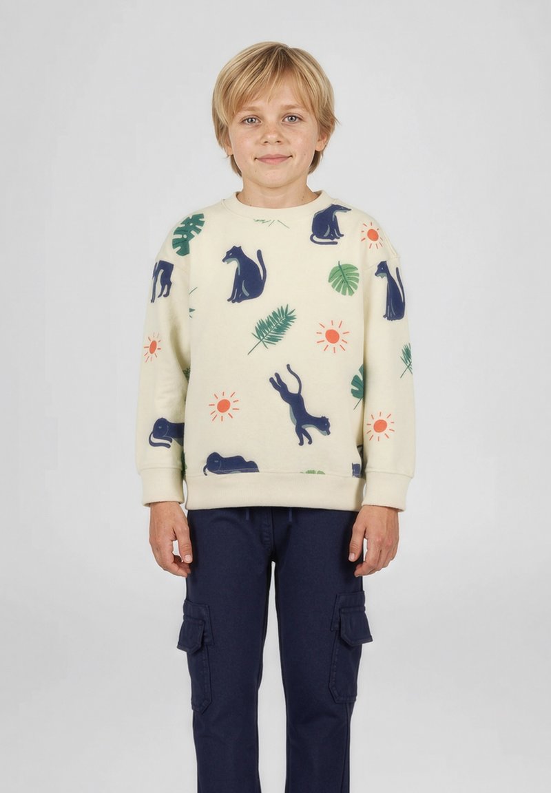 Dreng med blondt hår iført en cremefarvet sweatshirt med marineblå katte, grønne blade og orange sole, kombineret med marineblå cargobukser, stående på hvid baggrund.