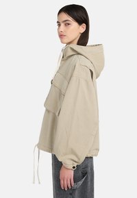 Jeune femme en veste beige à capuche avec poche avant et cordons, portant un pantalon en denim gris foncé, debout de profil sur fond blanc.