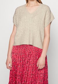 Haut en tricot beige à manches courtes avec un col en V et un motif en maille, associé à une jupe plissée rouge arborant un imprimé floral en noir et blanc.