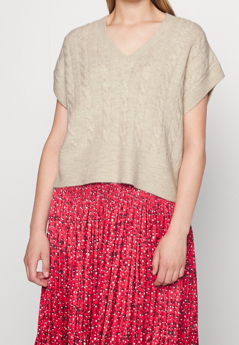 Haut en tricot beige à manches courtes avec un col en V et un motif en maille, associé à une jupe plissée rouge arborant un imprimé floral en noir et blanc.